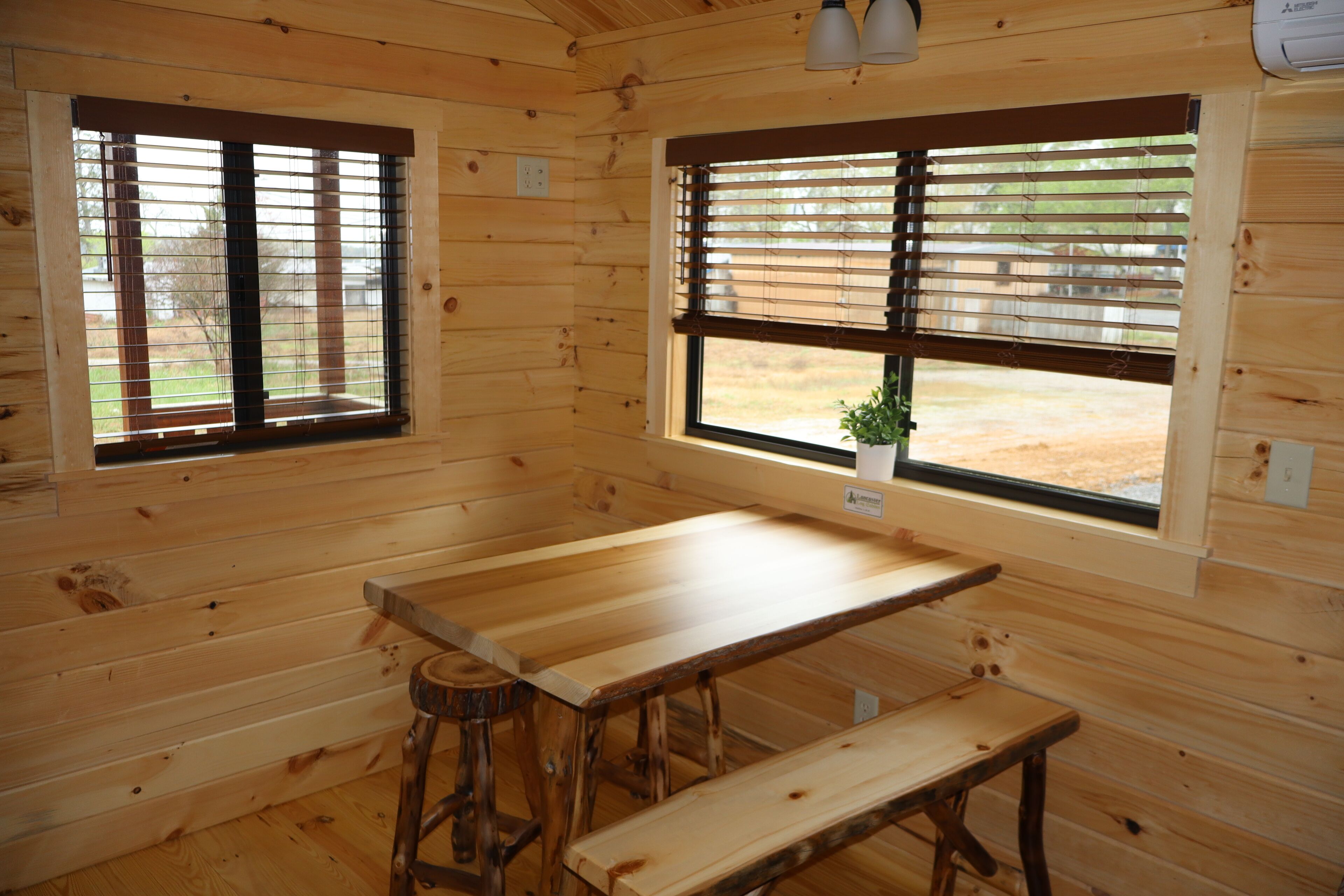 A wooden cabin interior with two windows, a wooden table, and a bench.