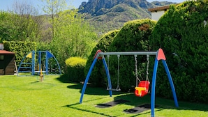 A playground with swings and a slide, surrounded by green grass and trees, with mountains in the background.