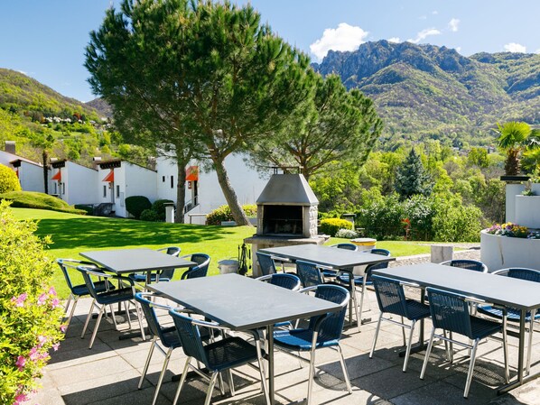 An outdoor patio with tables and chairs, a barbecue grill, and a view of mountains. - Villaggio Sureggio C10A by Interhome (Lugaggia)