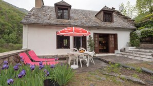Une maison avec un toit en tuiles, un balcon avec des fauteuils de jardin rouges, une table avec un parasol blanc et des fleurs violettes au premier plan.