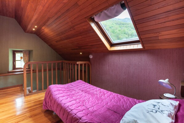 A bedroom with a sloped ceiling, a skylight, a bed with a pink bedspread, a nightstand with a lamp, and a window with curtains.
