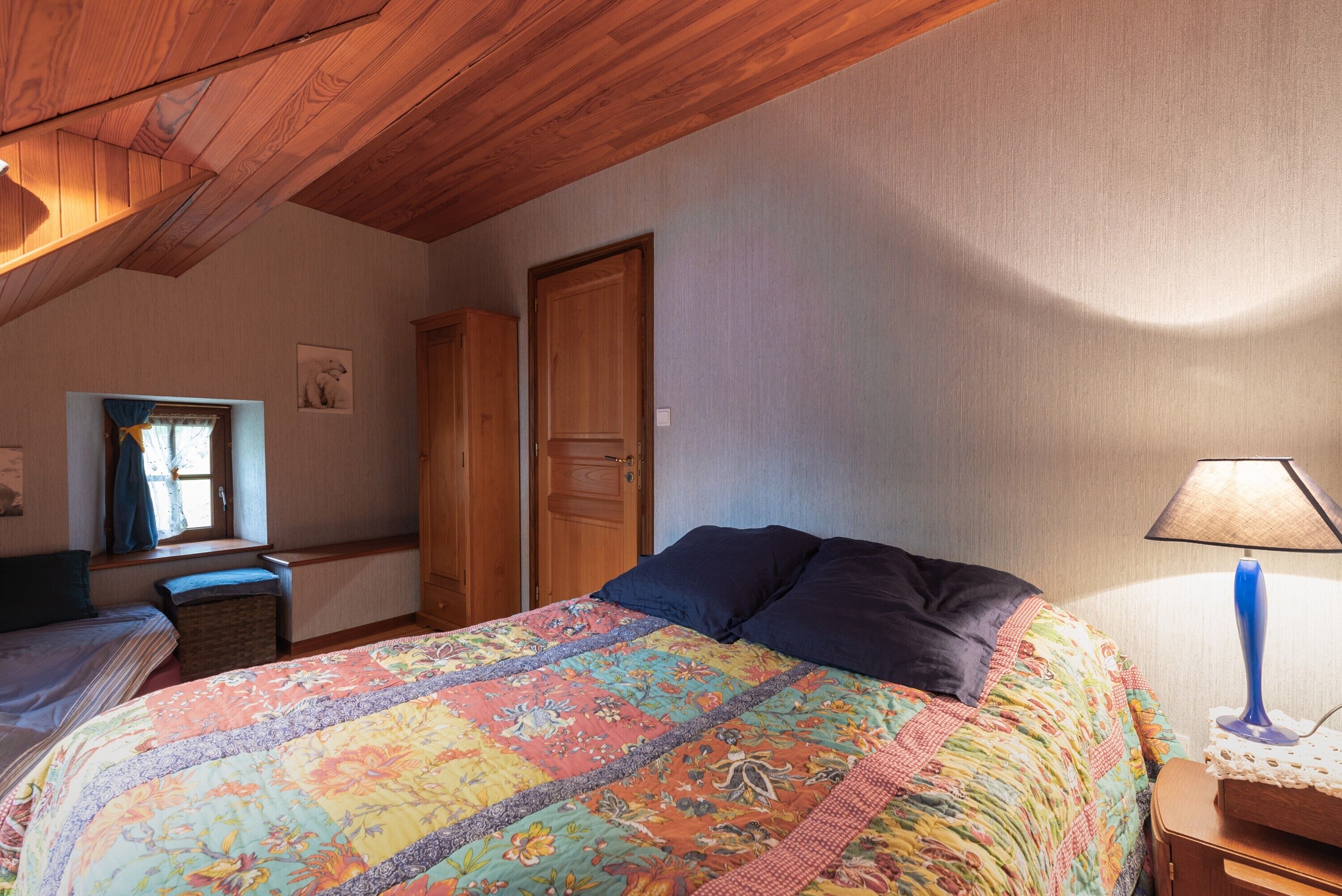 Un dormitorio con techo de madera, una cama con colcha floral, una mesita de noche con lámpara y una ventana con cortinas.
