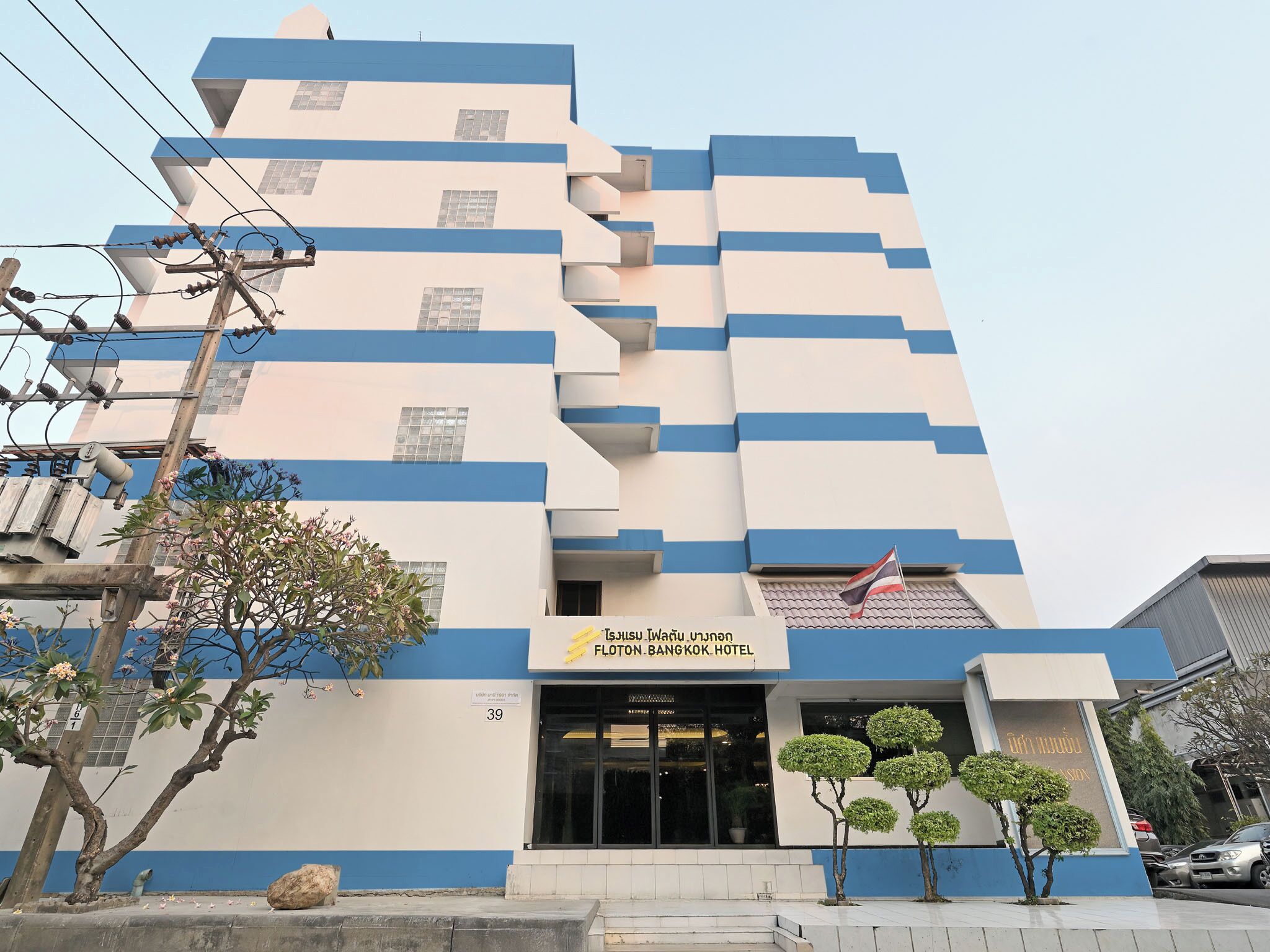 A modern hotel building with a blue and white facade, a sign indicating it's the Flotton Bangkok Hotel, and a flag on the roof.