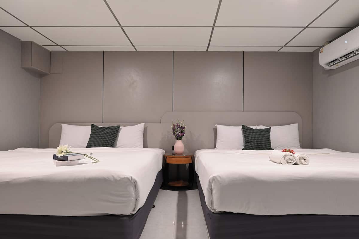 A hotel room with two beds, a nightstand, a vase with flowers, and a wall-mounted air conditioner.