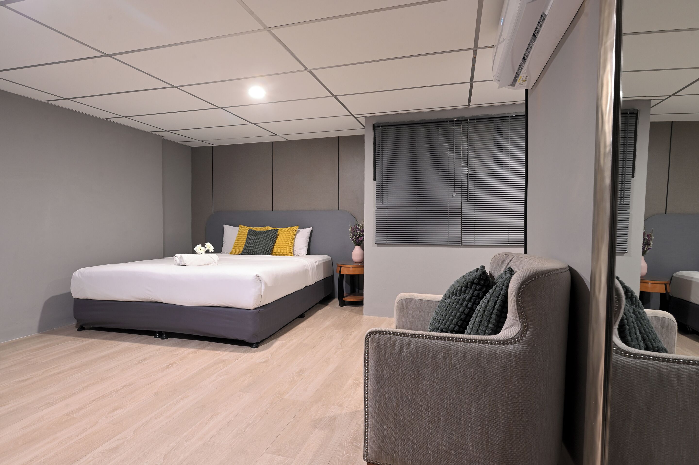 Une chambre d’hôtel moderne avec un grand lit, un fauteuil gris, une petite table d’appoint et une fenêtre avec des stores.