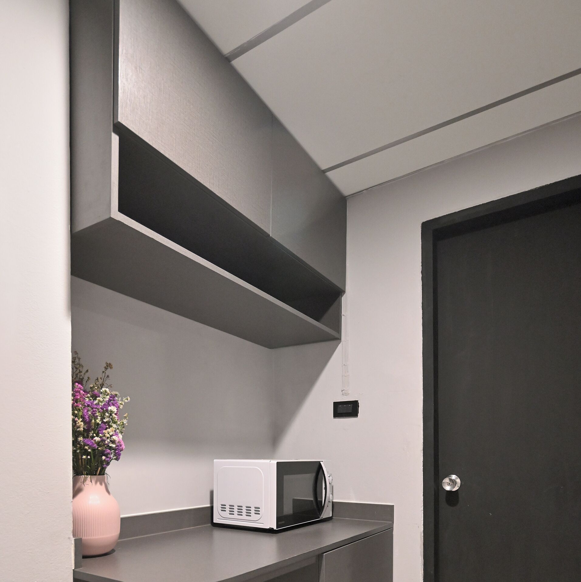 A modern kitchen with a microwave on a countertop, a pink vase with flowers, and a dark door.