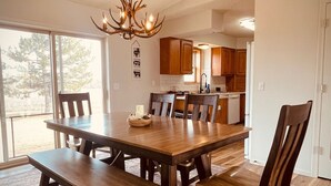 Un comedor amplio con una gran mesa de madera y bancos, una araña y vistas al exterior a través de grandes ventanales.