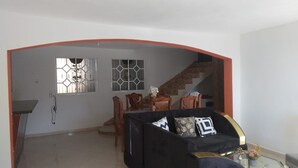 Ein Wohnzimmer mit Sofa, einem Esstisch mit Stühlen, einer Treppe und einem Fenster mit geometrischem Muster.