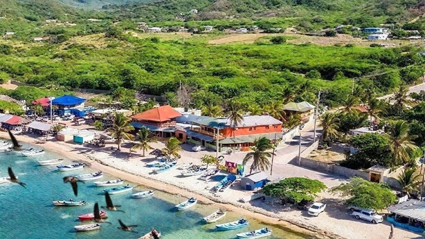 Eine Luftaufnahme einer Küstenlandschaft mit vertäuten Booten am Ufer, einem Strand und einer kleinen Stadt mit Gebäuden und viel Grün im Hintergrund.