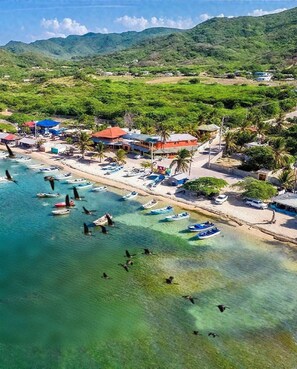 An aerial view of a coastal area with boats docked along the shore, a beach, and a small town with buildings and greenery in the background. - Hello Casa Mena Kompressa Mente Ekipa (Villa Vasquez)