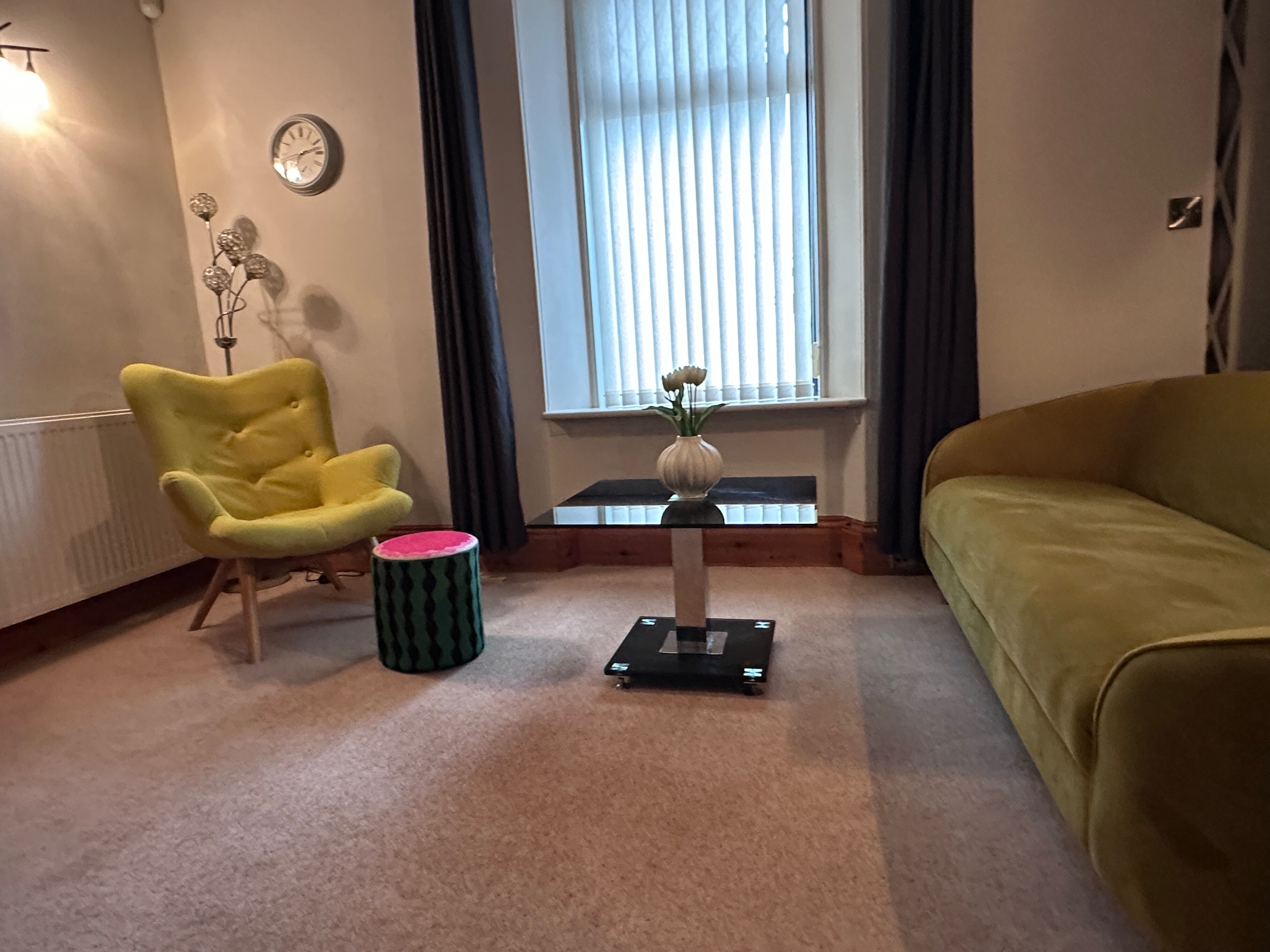 Una sala de estar con un sillón amarillo, un reposapiés verde y rosa, una mesita con un jarrón y un sofá verde.