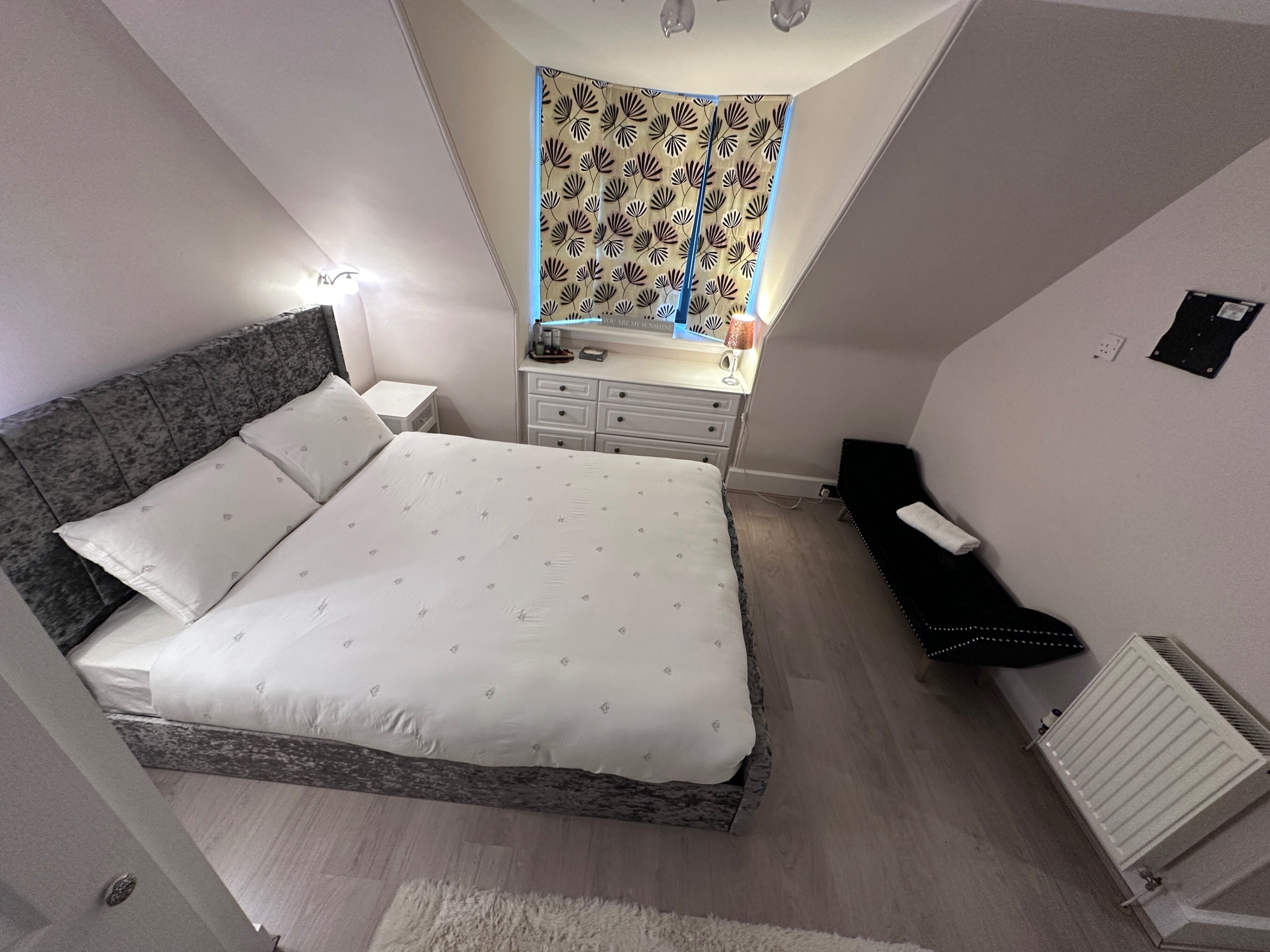 Een slaapkamer met een bed, nachtkastjes, een kaptafel, een radiator en een raam met een decoratief gordijn.