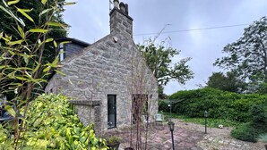 Una casa di pietra con un camino, una piccola finestra e un giardino con piante e un vialetto.