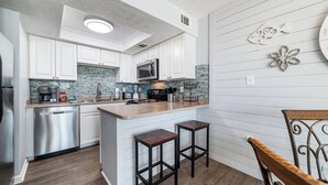 A modern kitchen with white cabinets, a stainless steel dishwasher, a microwave, and a tiled backsplash. There are two wooden stools and a small island with a sink. - Sunswept 705  Beach Front - King Bed -  POOL - Orange Beach  (Orange Beach)