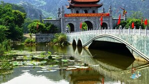 Un pont chinois traditionnel enjambant un étang où flottent des fleurs de lotus, le tout entouré d’une végétation luxuriante et de montagnes.