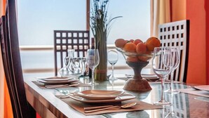 Una mesa de comedor dispuesta con platos, cubiertos y un bol de naranjas, con vista al mar al fondo.