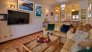 A living room with a flat-screen TV, a glass coffee table with fruit, a sofa with cushions, and a dining area in the background. - Ocean View-2 Bed-Sleeps 4-Pueblo Marinero (Acantilados de Los Gigantes)