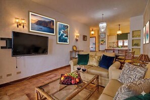 Una sala de estar con televisor de pantalla plana, una mesa de centro de vidrio con fruta, un sofá con cojines y una zona de comedor al fondo.