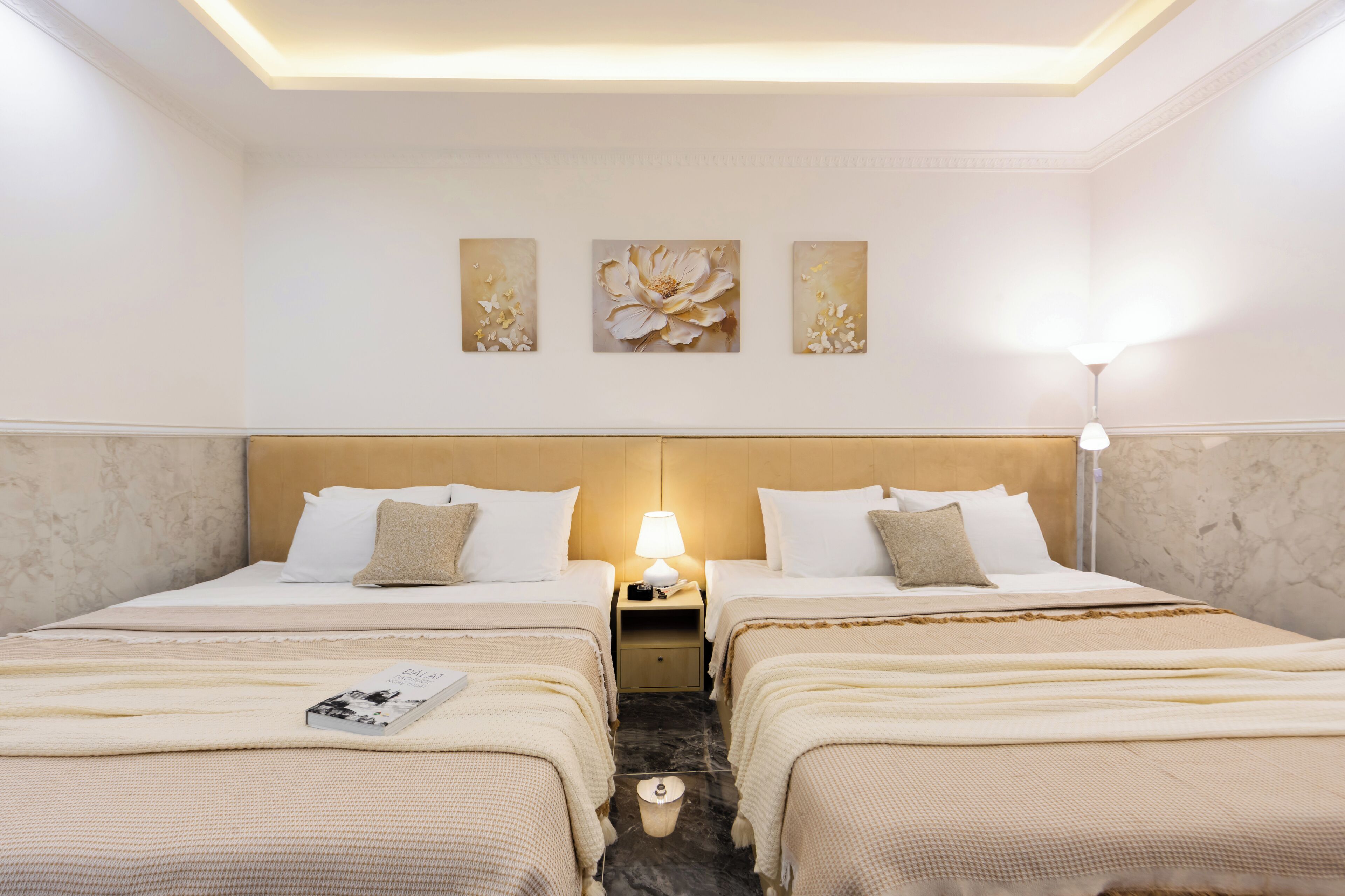 A hotel room with two beds, a nightstand, a lamp, and three floral artwork pieces on the wall.