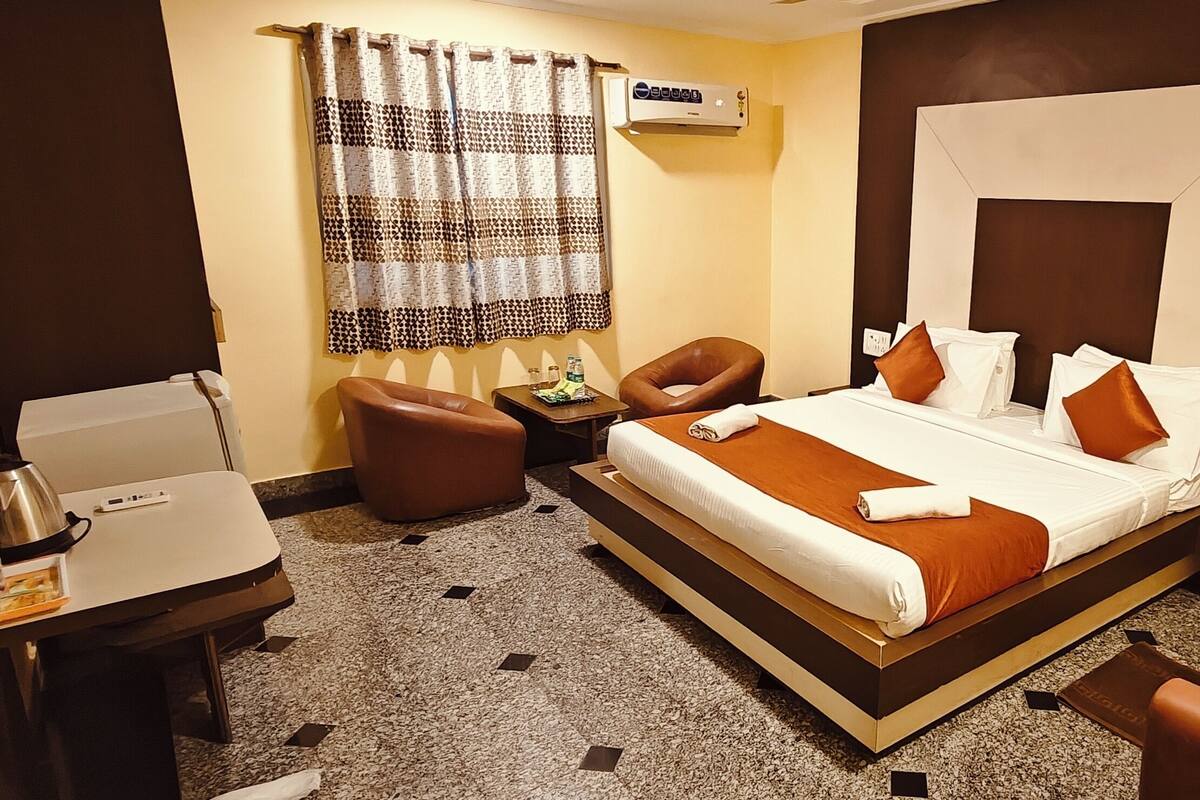Habitación de hotel con cama, dos sillas, una mesita con hervidor y botella, y aire acondicionado de pared.