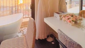 Quarto de hotel com varanda, banheira, cadeira de vime, escrivaninha com abajur e vista para o pôr do sol.