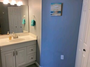 A bathroom with a white sink, mirror, and cabinets. There is a towel rack with two towels and a framed quote on the wall. - Beach Condo • Walk to Sand • 2 Pools • Balcony • Free Beach Gear (Panama City)