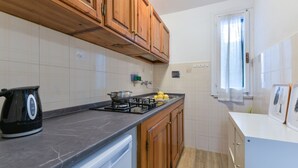 Une cuisine avec des meubles de rangement en bois, une cuisinière à gaz, une bouilloire et une fenêtre avec des rideaux.