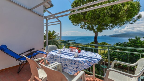 Een balkon met een tafel gedekt voor twee personen, met uitzicht op de kust, een boom en een heldere lucht.