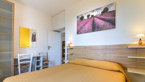 Een slaapkamer met een bed, een bureau en een stoel. Er hangt een schilderij aan de muur en op het nachtkastje staat een lamp.