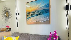 Ein modernes Wohnzimmer mit einem grauen Sofa, einem runden Couchtisch, einem kleinen Beistelltisch, einem Gemälde mit Strandmotiv und einem Wanddekor.