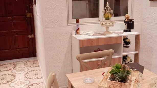 A dining area with a wooden table, chairs, a window, and a decorative shelf with plants.