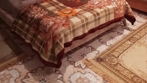 A bedroom with a large bed, a headboard, a floral bedspread, a nightstand, a vase with flowers, and a framed picture on the wall.