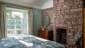 Ein Schlafzimmer mit großem Fenster, Kamin aus Stein, Holzkommode und floralem Bettbezug.