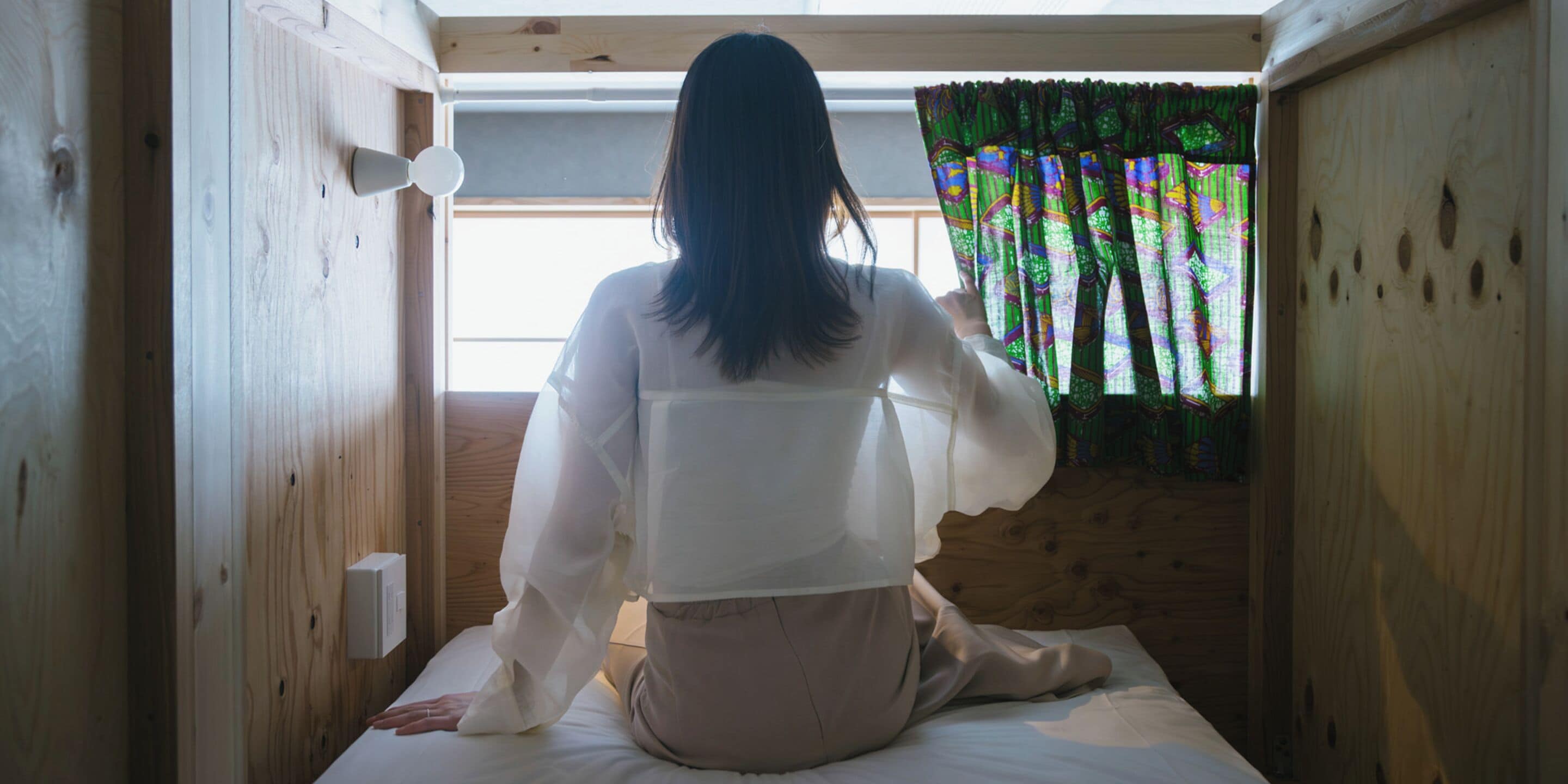 Una persona sentada en una cama en una habitación con paredes de madera y un ventanal con cortinas de colores.