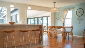 Eine moderne Küche mit Holzbarhocker, einem Esstisch und Stühlen. Große Fenster sorgen für natürliches Licht und einen Ausblick ins Freie.