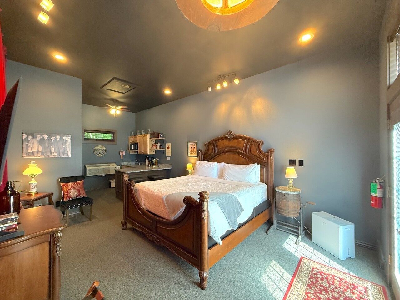 A bedroom with a wooden bed, a desk, a chair, a radiator, a small table with a lamp, and a window with blinds.