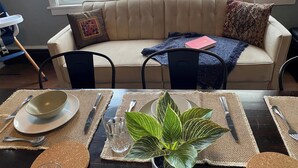 Un comedor con una mesa dispuesta para dos, un sofá color beige con cojines estampados y una ventana con cortinas traslúcidas.