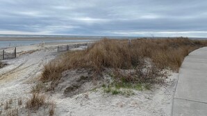 Una playa con un camino de arena que lleva al agua, una valla y una duna cubierta de hierba.