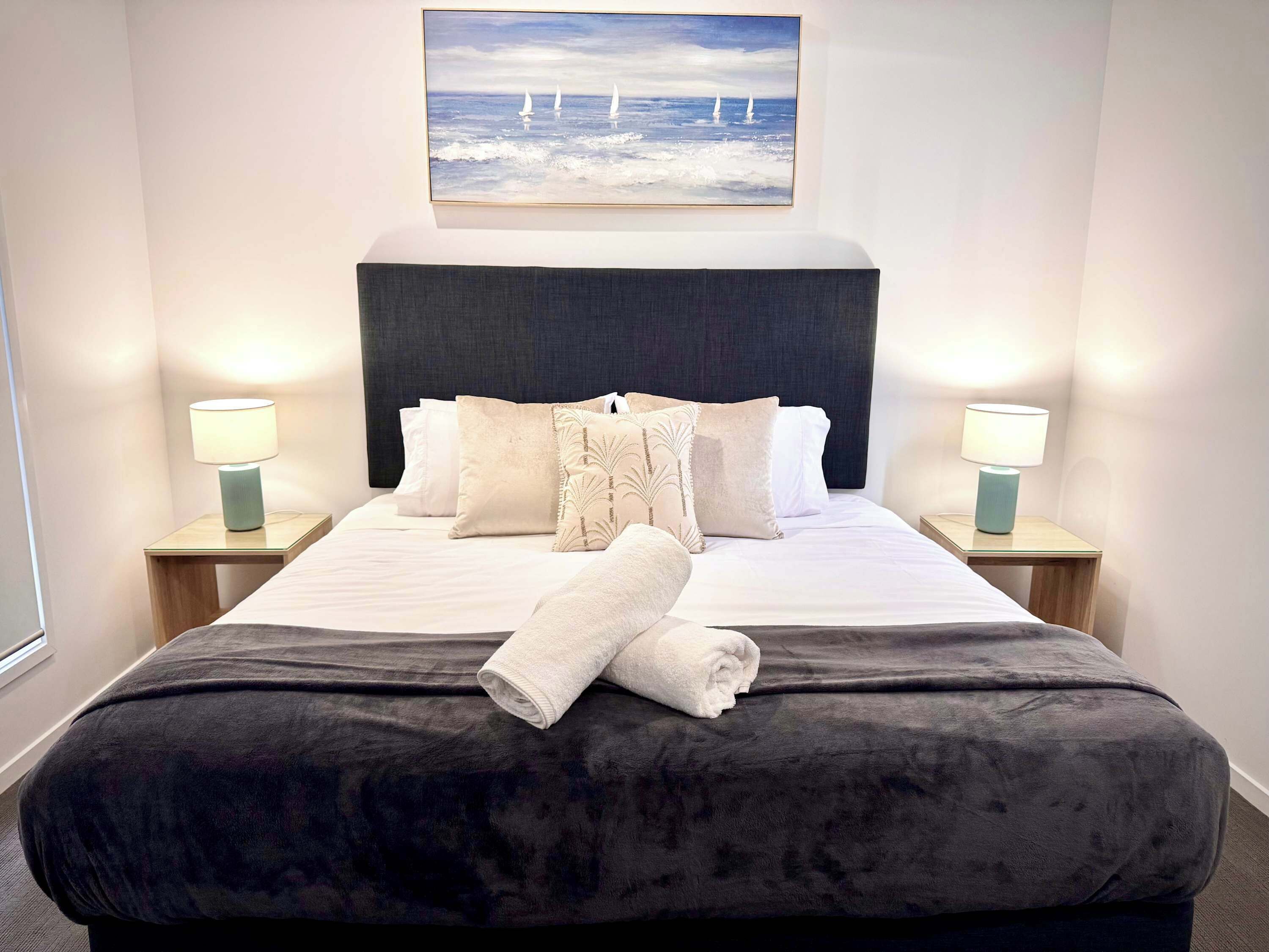A neatly made bed with a dark headboard, two bedside lamps, and a painting of sailboats on the wall.