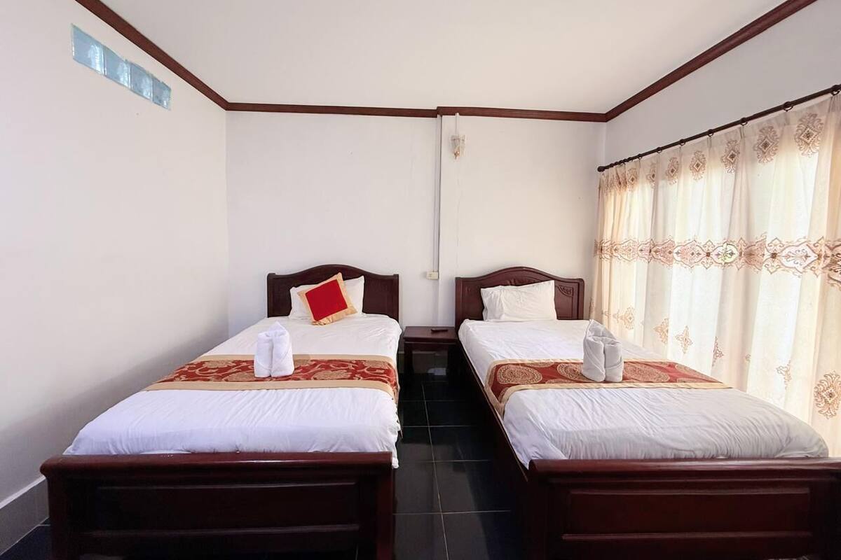 Een hotelkamer met twee enkele bedden, elk met een rood-wit kussen en een rode en gouden sprei. Aan de rechterkant is een raam met gordijnen.