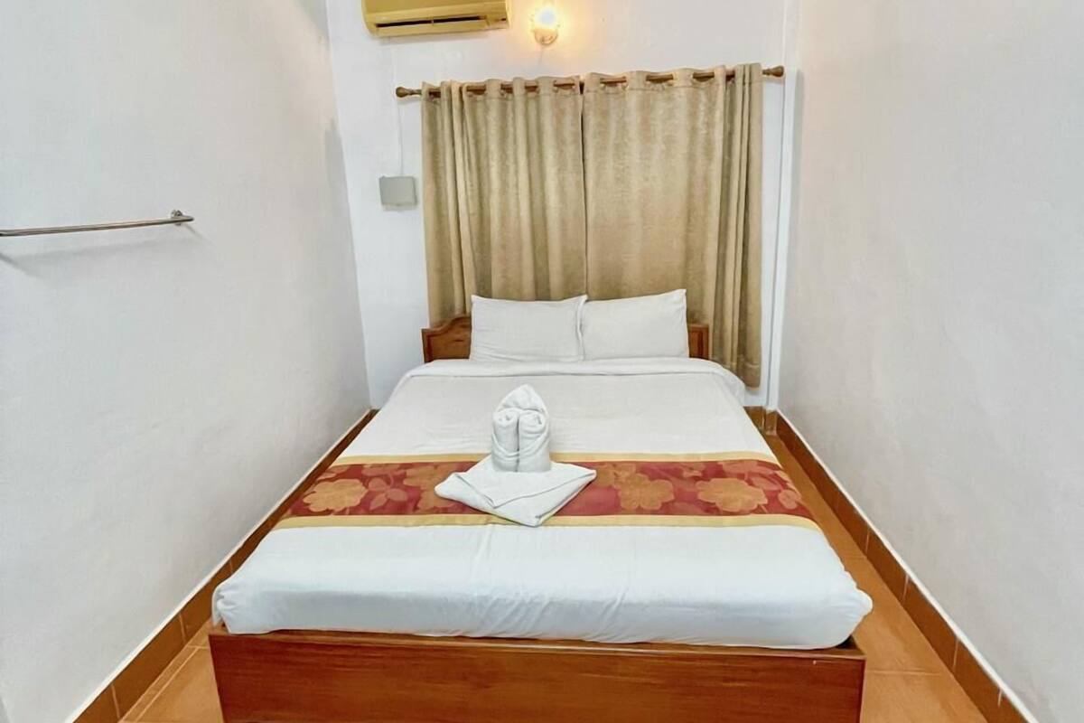 Een hotelkamer met een enkel bed, een houten hoofdbord, een nachtkastje en een wandairconditioner.