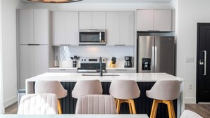 A modern kitchen with white cabinetry, a stainless steel refrigerator, a microwave, and a coffee machine. There are four bar stools and a white dining table. - Perfect Family Getaway, 4BR & Themed Magic (Kissimmee)