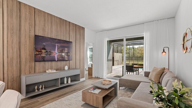 A modern living room with a large flat-screen TV mounted on a wooden wall, a grey sofa, a coffee table, and a sliding glass door leading to an outdoor area. - Private Pool Home Near Disney & Themed Rooms (Kissimmee)