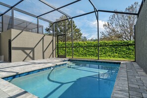 Ein Schwimmbecken mit Glasüberdachung, umgeben von einer Hecke und einem gepflasterten Weg.
