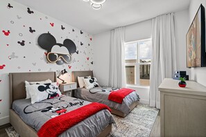 Ein Hotelzimmer mit zwei Betten, Mickey Mouse Wandbildern, einem Fenster mit Vorhängen, einem Schreibtisch mit Pflanze und einem Fernseher an der Wand.