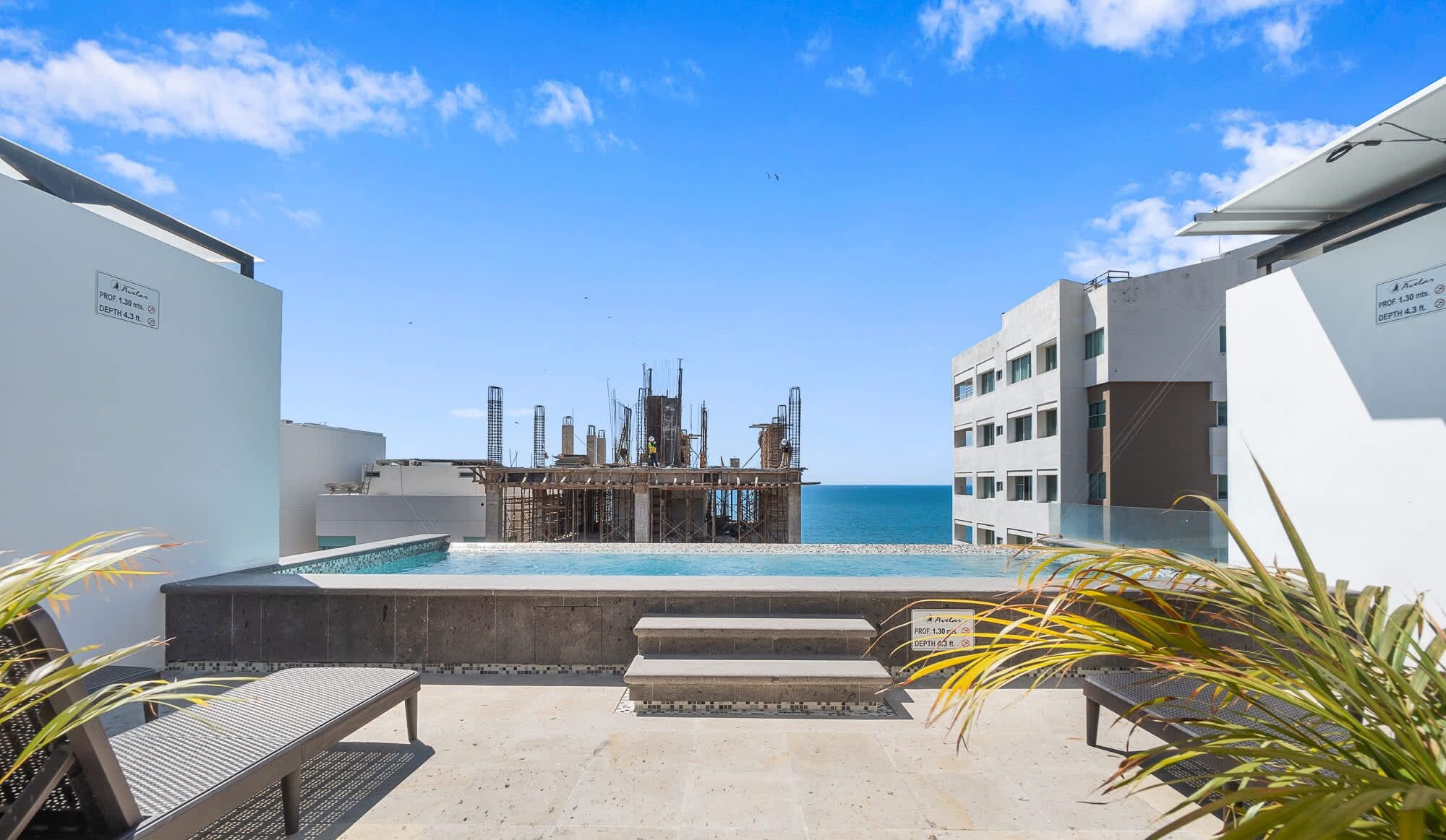 A pool area with a view of the sea, a construction site in the background, and a building with a sign indicating a depth of 4.3 meters.