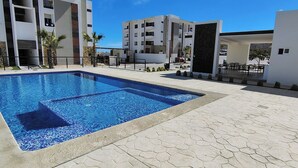 A swimming pool with a tiled edge, surrounded by a paved area with a modern building in the background. - Charming 2-bedroom apartment in Los Cabos with WiFi, AC, Swimming Pool and more. (B.C.S.)