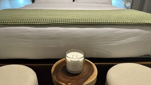 Una cama bien tendida con una manta verde, una vela encendida sobre una mesa de madera y dos sillones acolchados.