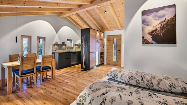Een ruime kamer met een houten plafond, een bed met bloemenbedrukking, een zithoek met een houten tafel en stoelen, en een keukenhoek.