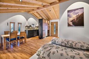 Une pièce spacieuse avec un plafond en bois, un lit recouvert d’une literie à motifs floraux, un coin repas avec une table et des chaises en bois, et un coin cuisine.